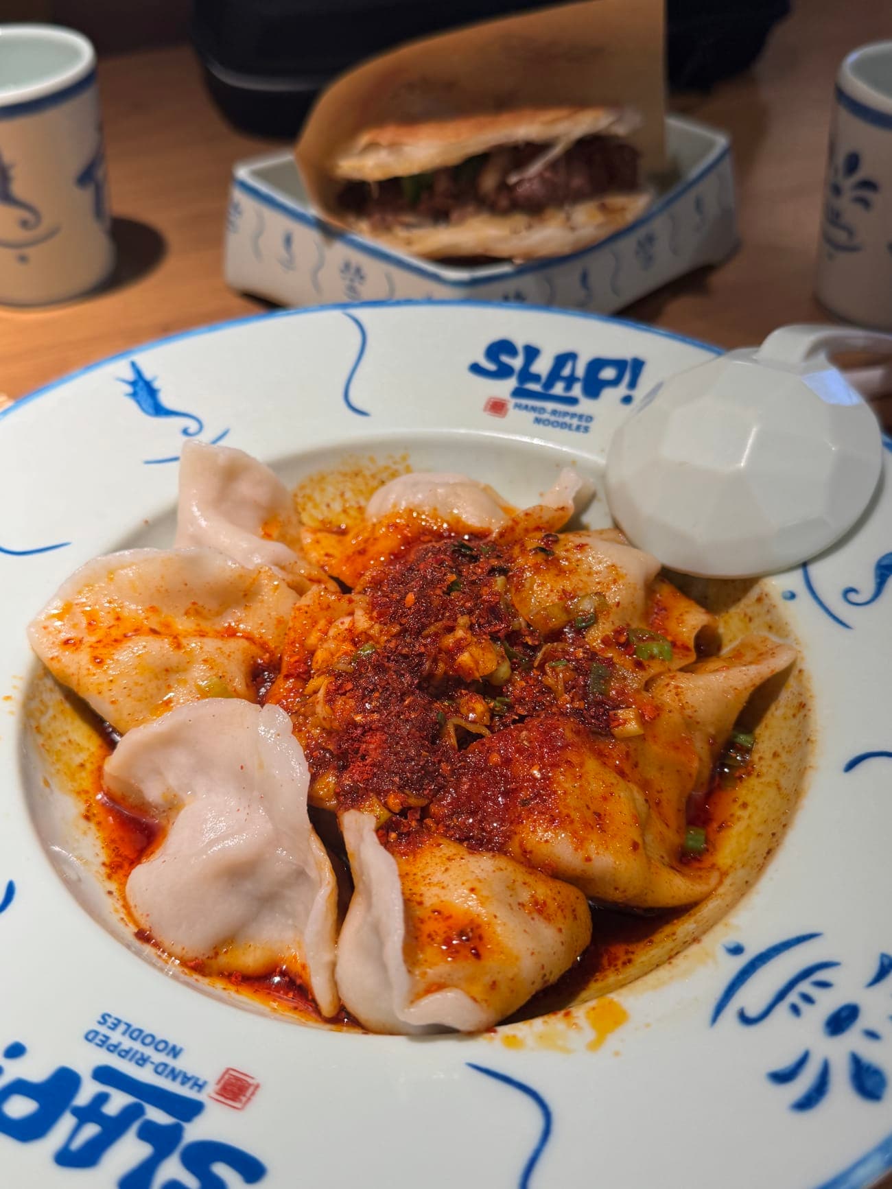 Plate of ten plump beef onion dumplings smothered in red chili oil with chopped scallions at SLAP Hand-Ripped Noodles