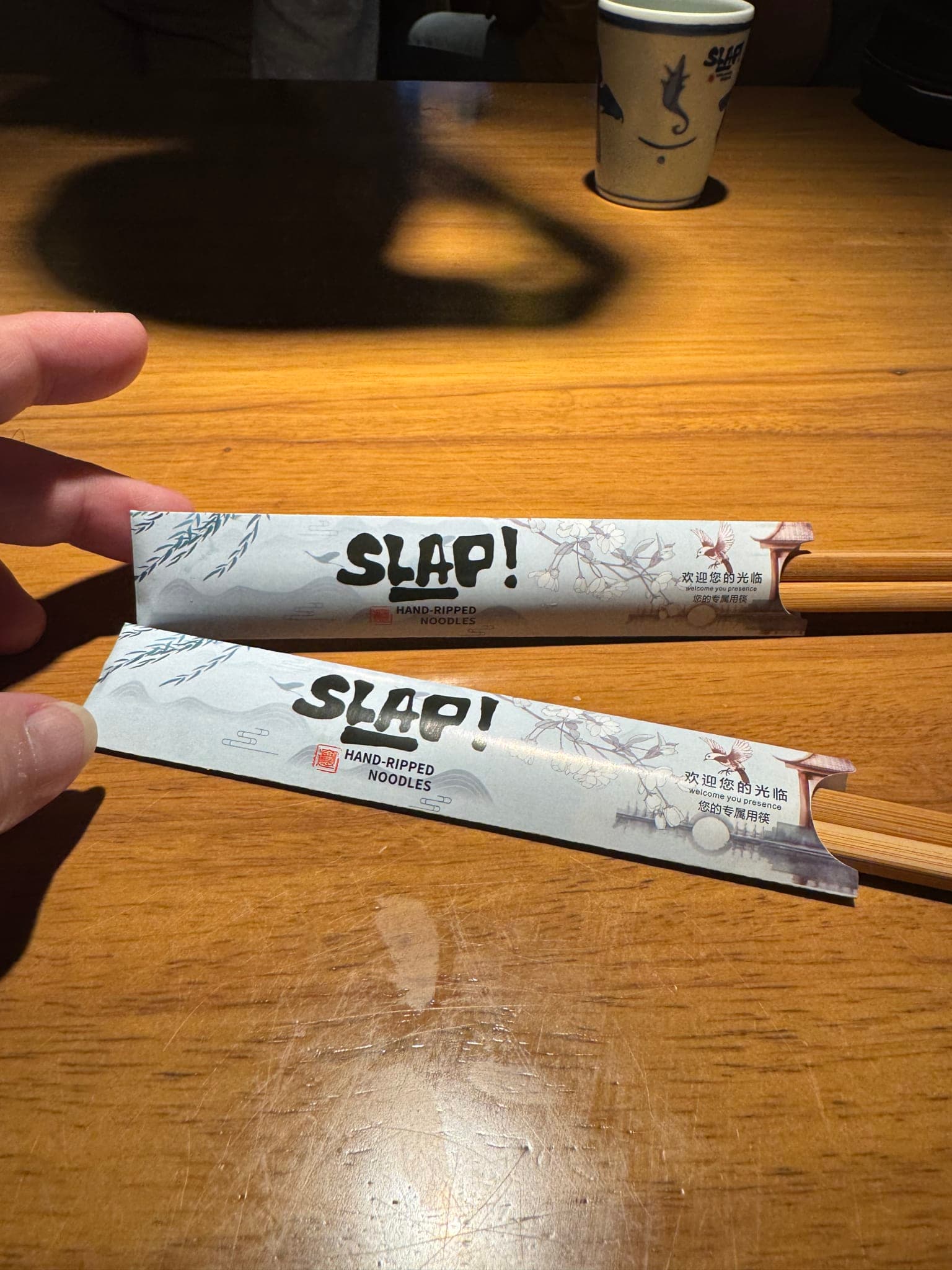 Two pairs of SLAP-branded chopsticks with floral paper sleeves on a wood table at SLAP Hand-Ripped Noodles