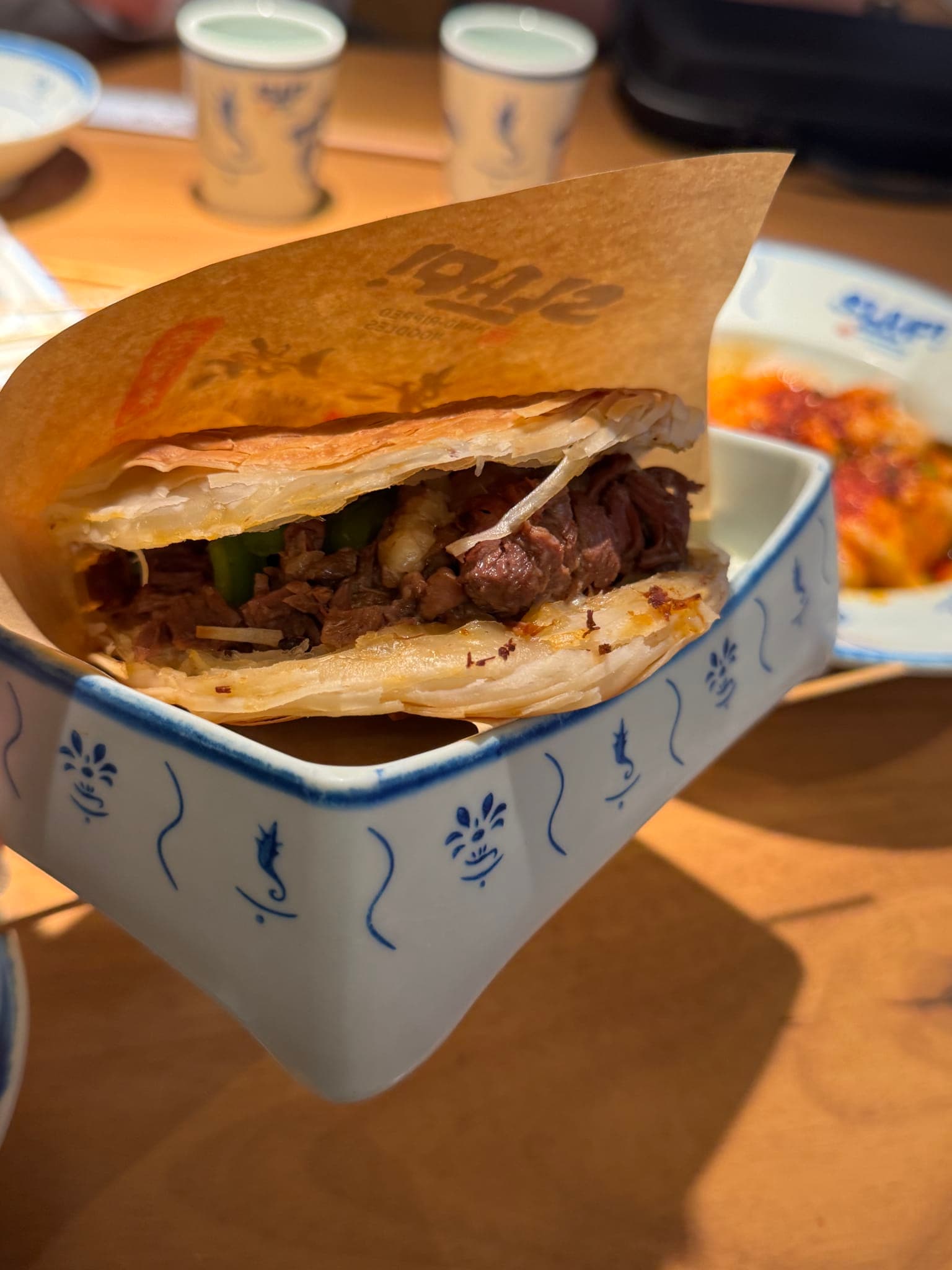 Flaky crispy pancake stuffed with braised beef and green peppers wrapped in parchment in a SLAP-branded ceramic dish