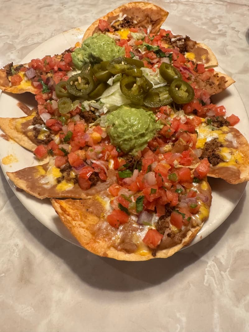 A large oval plate of Chuy's Panchos loaded with seasoned ground beef, refried beans, and melted cheese on crispy tostada chips, topped with shredded lettuce, fresh pico de gallo, sliced pickled jalapeños, and a scoop of guacamole