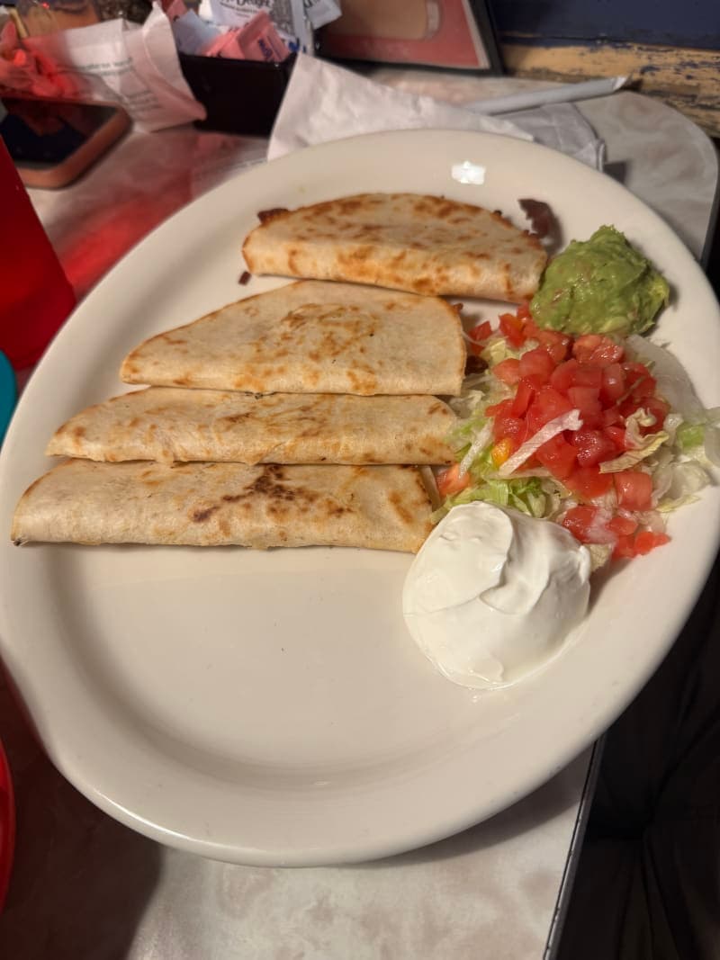 A golden, grilled flour tortilla quesadilla cut into wedges and filled with melted cheese, served on a plate with a small bowl of sour cream and a small bowl of fresh guacamole alongside