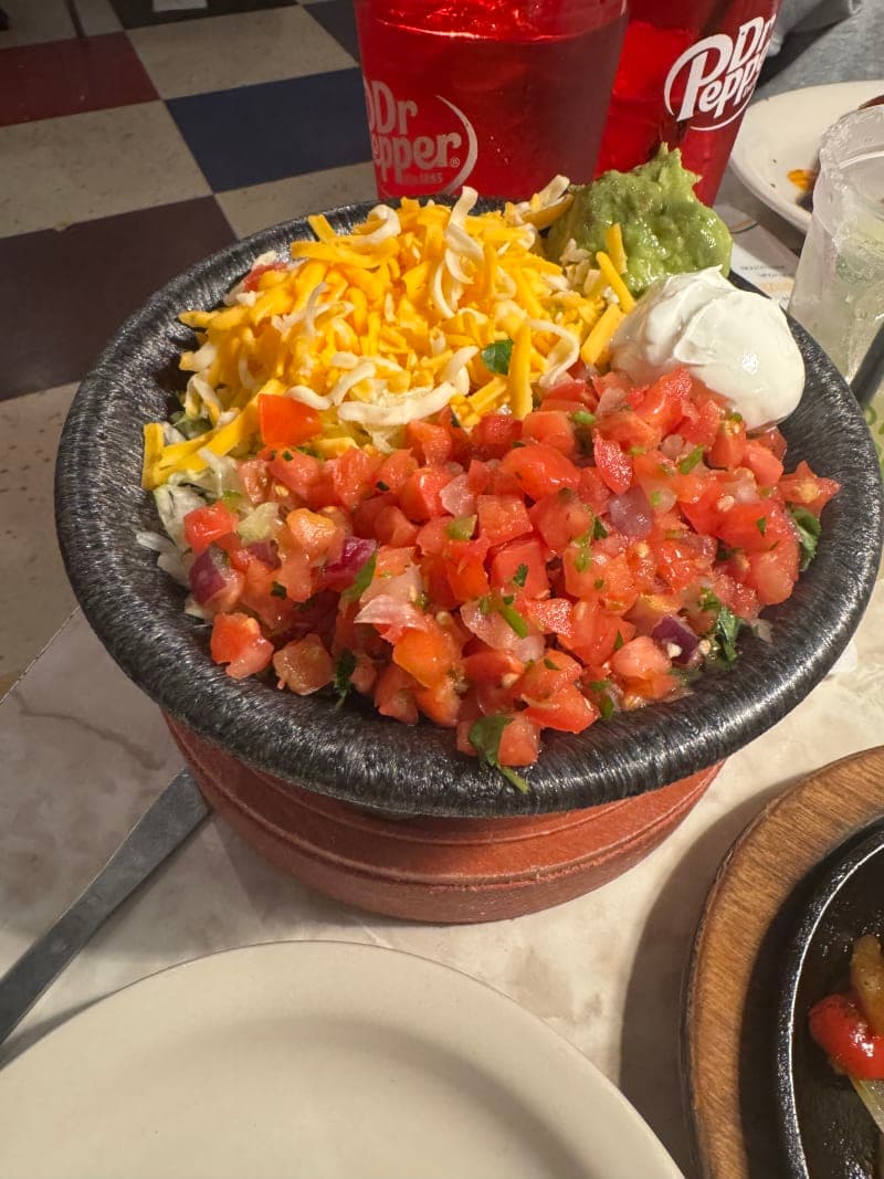 A large bowl filled with fresh pico de gallo, shredded cheese, chunky guacamole, and sour cream served together as a single fajita topping dish